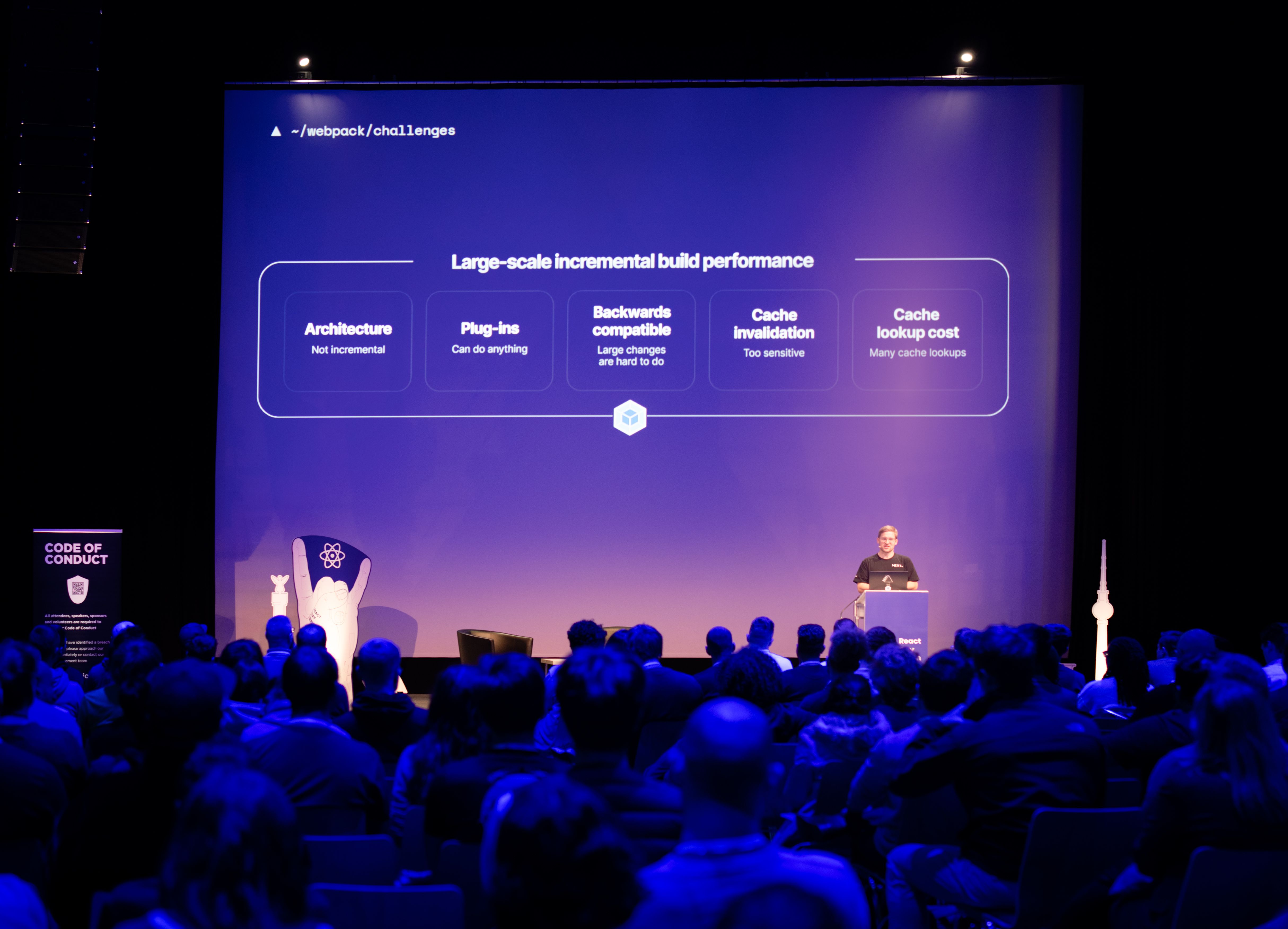 Salle avec vue sur l'écran d'information, le présentateur avec un t-shirt noir à gauche sur scène prenant la parole.